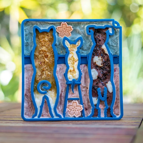 A blue silicone SodaPup Waiting Dogs Design ETray Enrichment Tray for Dogs, shaped like a cat, bunny, and dog, sits on wood with colorful mixtures, serving as a slow feeder or lickmat against a blurred outdoor backdrop.