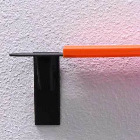 A black wall-mounted holder secures an orange pen against a white textured wall, resembling Chatelet Manufacturing’s Toy Racetrack Wall Connectors Compatible With Hot Wheels, with a faint pinkish-red reflection nearby.
