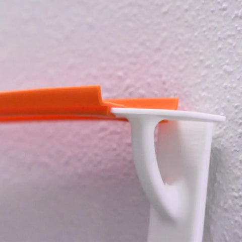 Close-up of an orange Chatelet Manufacturing Toy Racetrack Wall Connector for Hot Wheels, resting on a white bracket with a curved support, attached to a light-textured wall. Made in USA.