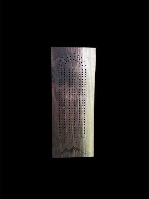 The Walnut Live Edge Cribbage Board by B. James Design & Woodwork features multiple rows of holes and a subtle engraved mountain at the bottom, all handcrafted and shown against a black background.
