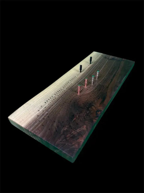 The Walnut Live Edge Cribbage Board by B. James Design & Woodwork features handcrafted quality, a resin inlay, colored pegs, and built-in peg storage, shown against a striking black background.