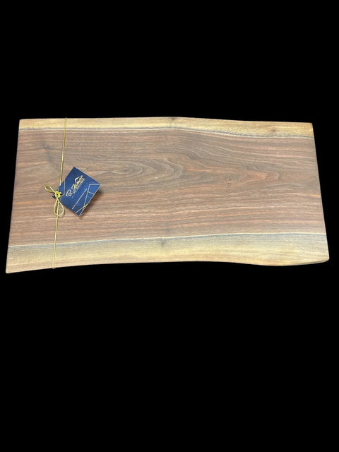 The Walnut Live Edge Serving Tray by B. James Design & Woodwork is a handcrafted rectangular walnut board with a natural live edge and a brown tag tied with yellow string on one corner, shown against a black background.