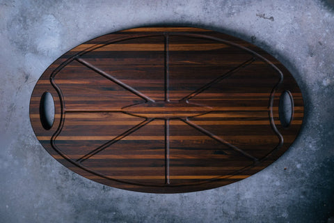 The Oval Carver - Walnut by Montana Block is an oval walnut cutting board with dark and light stripes, deep grooves, and dual handles, handcrafted in the USA and shown on a textured gray surface.