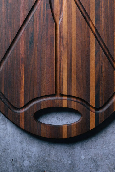 A close-up of Montana Blocks The Oval Carver - Walnut, a handcrafted USA-made cutting board with a striped pattern and oval handle, placed on a gray textured surface.
