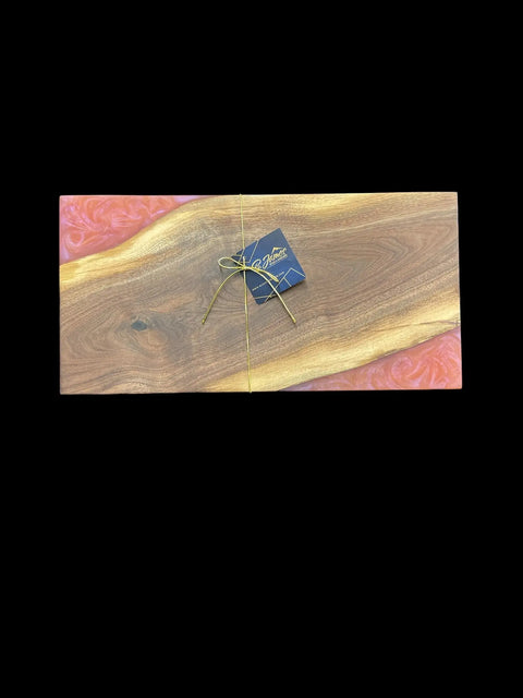 The Sunset Epoxy Walnut Serving Tray by B. James Design & Woodwork is handcrafted in the USA, featuring pink resin-edged walnut, tied with gold string and a blue tag, displayed on a black background.