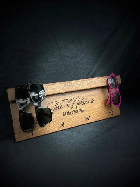 The B. James Design & Woodwork Sunglasses and Key Rack - Walnut holds three pairs of sunglasses with hooks, ideal for entryway organization, and features The Nelsons Est. March 22nd, 2014 elegantly engraved on the front.