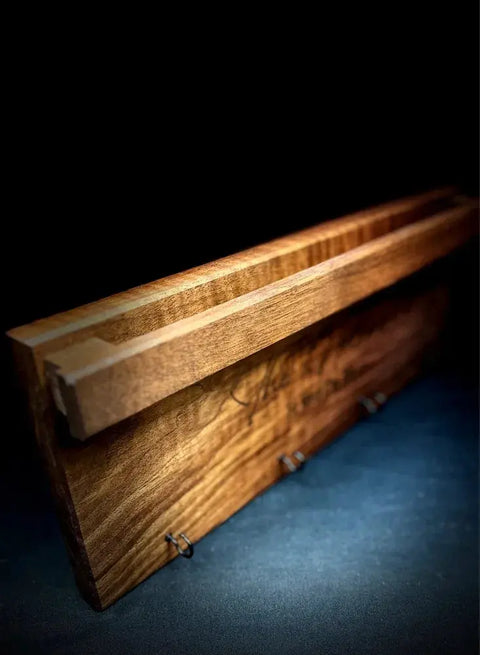 A close-up of the Sunglasses and Key Rack - Walnut by B. James Design & Woodwork, featuring a handcrafted walnut shelf with a groove for sunglasses storage and three front metal hooks for keys, ideal for entryway organization against a dark backdrop.