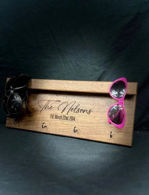The B. James Design & Woodwork Sunglasses and Key Rack - Walnut, engraved with “The Nelsons. Est. March 22nd, 2014.”, holds three sunglasses (black and pink shown) for a stylish, organized entryway.