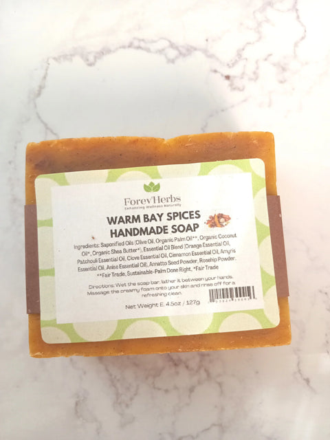 A rectangular bar of Warm Bay Spices Handmade Soap by Forev'Herbs rests on a light surface. The label features organic oils among its natural ingredients and highlights that the soap is handmade, fair trade, and sustainably crafted.