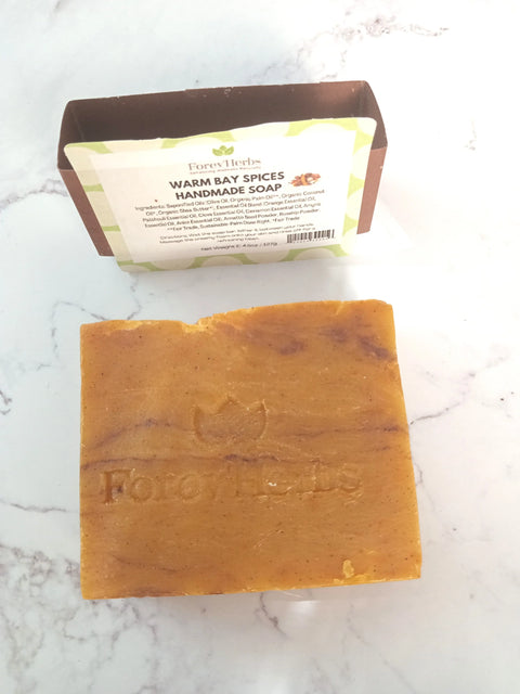 An orange-brown rectangular bar of Warm Bay Spices Handmade Soap from Forev'Herbs rests on white marble, with its labeled packaging displaying organic oils and detailed ingredients behind it.