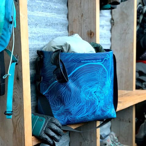 The eqpd GearBucket - Washington Pass, a blue tote with a felt base and topographic map design, sits on a wooden shelf filled with outdoor gear like a jacket and gloves, alongside other bags and equipment.