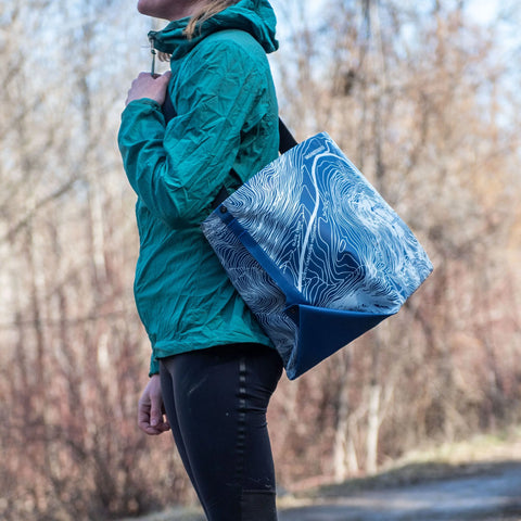 Wearing a teal jacket and black leggings, a person stands outdoors with the eqpd GearBucket - Washington Pass, a spacious blue and white tote bag featuring a felt base and topographic map design, slung over their shoulder.