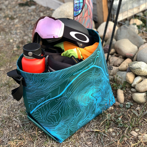 The eqpd GearBucket - Washington Pass, featuring a topographic map design and felt base, sits on rocky ground filled with a red water bottle, black helmet with pink goggles, and outdoor gear.