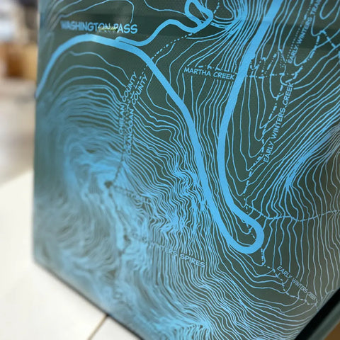 A close-up of the eqpd GearBucket - Washington Pass, a blue topographic map with a felt base highlighting Washington Pass, Okanogan County, Martha Creek, Early Winters Creek, Spires, and elevation contour lines.
