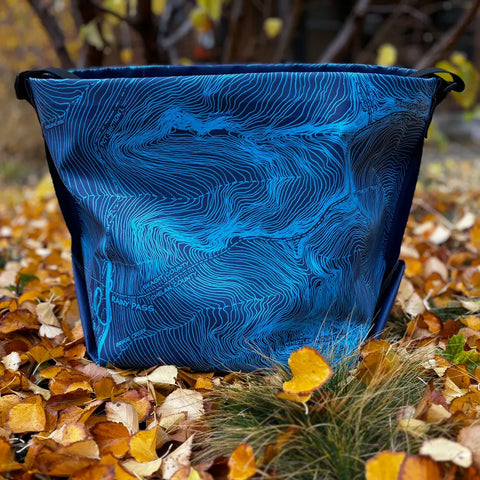 The eqpd GearBucket - Washington Pass, a blue tote with topographic lines and a felt base, rests on autumn leaves with blurred branches and foliage in the background.