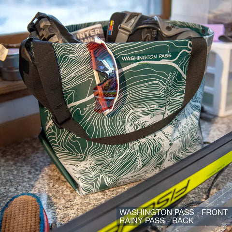 The eqpd GearBucket - Washington Pass is a green and white tote bag with a felt base and topographic map design, shown on a counter holding sunglasses, gloves, and gear. Text: WASHINGTON PASS - FRONT, RAINY PASS - BACK.