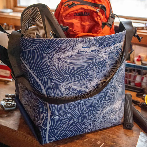 The eqpd GearBucket - Washington Pass, featuring a blue and white topographic map design with a felt base, holds an orange backpack and outdoor gear on a cluttered wooden table near a window, with a plastic storage bin nearby.