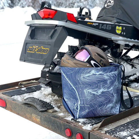 A snowmobile sits on a snowy trailer, with a brown helmet, pink goggles, and the eqpd GearBucket - Washington Pass tote bag nearby. Snow-covered trees create a blurred background.