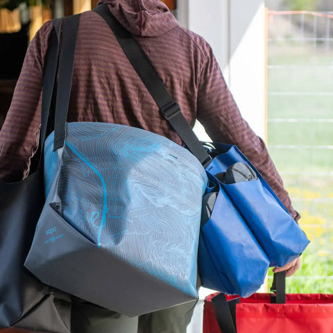 A person in a brown striped hoodie carries multiple eqpd GearBucket - Washington Pass totes in blue, light blue, black, and red while walking through an open door.