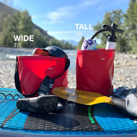 Two red eqpd GearBucket waterproof bags with watertight seams, labeled WIDE 27L and TALL 18L, sit on a paddleboard by a river with water shoes, paddle, helmet, pump, and gear amid trees and mountains.