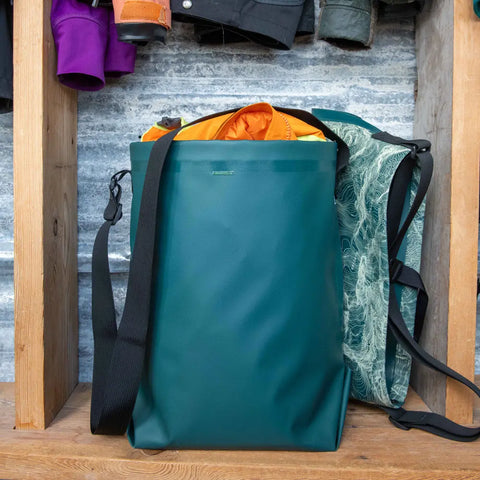 An eqpd GearBucket, a teal waterproof tote with watertight seams and black strap, sits on a wooden shelf. Bright orange fabric peeks out and a green patterned bag is beside it. Coats and clothing hang above.