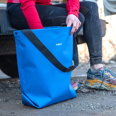 A person in athletic wear and worn running shoes sits on a vehicles edge, holding an eqpd GearBucket tote with watertight seams, outdoors on a dirt surface.