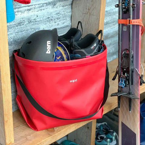 A red eqpd GearBucket filled with a black helmet and ski gear sits on a wooden shelf beside leaning skis, a plastic storage bin, and other outdoor equipment.