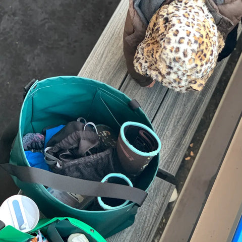 Overhead view of a child in a leopard-print hat leaning over a bench next to an eqpd GearBucket tote in teal, filled with boots, clothes, and other items outdoors.