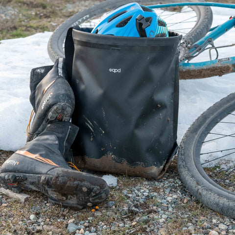 A pair of muddy cycling boots, an eqpd GearBucket (black, waterproof with watertight seams), and a blue helmet rest on the ground beside a bicycle on gravel and snow.