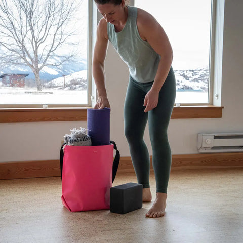 A woman in workout clothes stands indoors near a window with snowy mountains outside, placing a rolled yoga mat into an eqpd GearBucket tote. A yoga block and blanket rest on the floor.