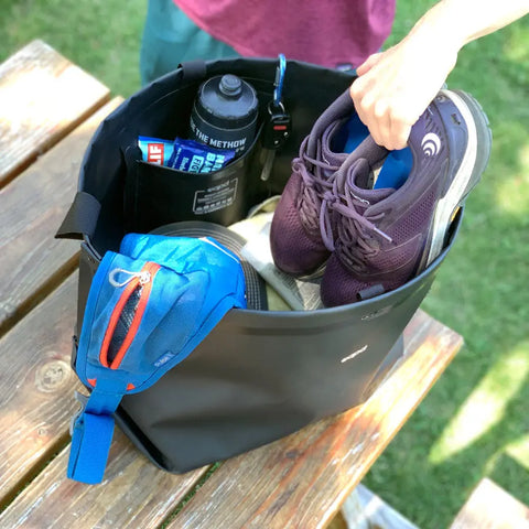 A hand places purple running shoes into a black eqpd GearBucket on a wooden picnic table. Inside the GearBucket are a blue pouch, water bottle, and CLIF bar, with grass visible in the background.