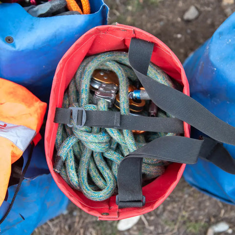 Top view of a red eqpd GearBucket holding a coiled climbing rope and metal carabiners, surrounded by blue bags on the ground.