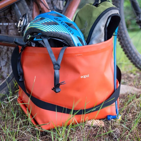 An orange eqpd GearBucket sits on the grass by a tree and bike, holding a blue and black helmet and a green backpack. Its watertight seams protect your gear better than any plastic storage bin.