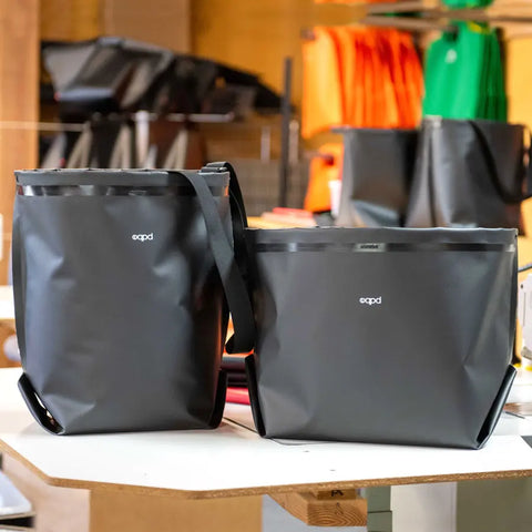 Two black eqpd GearBucket tote bags sit on a table in a workshop, with brightly colored bags like orange and green blurred in the background, evoking the atmosphere of a bag-making or design studio.