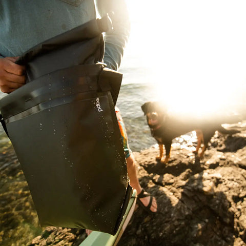 A person stands by the water with a black eqpd GearBucket - RollTop leakproof dry bag under bright sunlight, while a dog is on the rocky shore in the background.