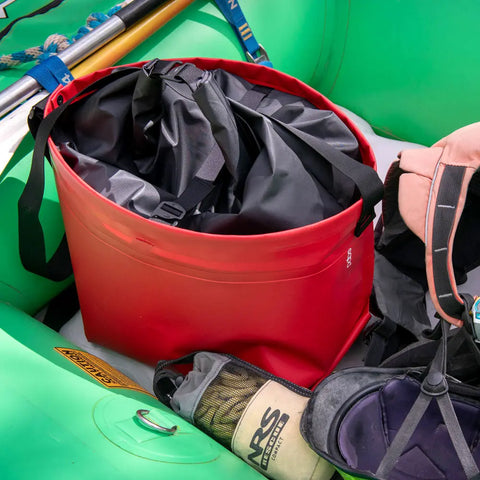 The eqpd GearBucket - RollTop red leakproof dry bag sits open in a green inflatable raft beside a beige water bottle, black sandal, and peach life jacket, revealing its black inner material and straps.