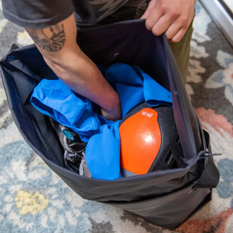 A person with a tattoo reaches into a black eqpd GearBucket - RollTop holding a blue jacket, orange helmet, and climbing gear. The watertight tote rests on a patterned rug.