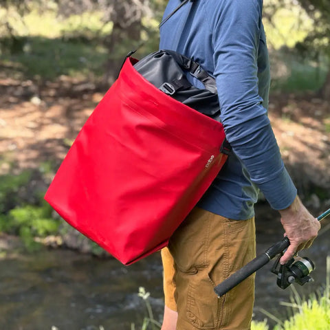 Outdoors, someone holds a fishing rod and carries an eqpd GearBucket - RollTop, a large red watertight tote, over their shoulder with trees and a stream in the background.
