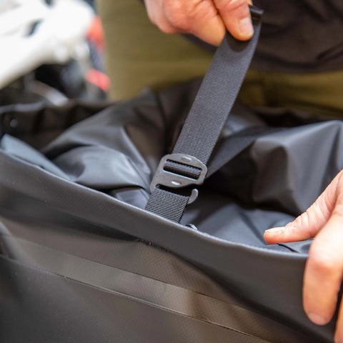 A person’s hands fasten a black plastic buckle on an eqpd GearBucket - RollTop dry bag, securely closing the top to keep contents protected.