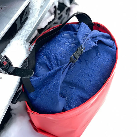 A blue eqpd GearBucket - RollTop dry bag, leakproof and covered in droplets, is secured inside a red container mounted on a snow-covered vehicle, likely a snowmobile.