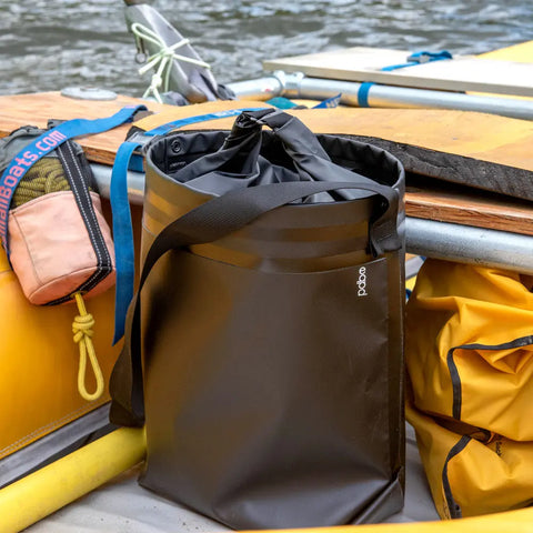 A black eqpd GearBucket - RollTop dry bag with a shoulder strap sits on a raft, surrounded by yellow bags, paddles, and river GearBuckets on a wooden platform near the water.