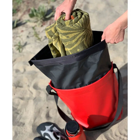 A person places a rolled green towel into the eqpd GearBucket - RollTop dry bag on sandy ground, with water shoes and a red bottle nearby. Small plants are visible in the sand.