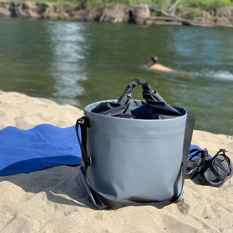 An eqpd GearBucket - RollTop gray leakproof dry bag rests on a sandy beach near a blue towel and sandals, while someone swims in the river with lush greenery across the bank.