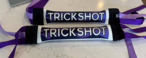 Two rectangular Dock Diving Bumper Tug toys (Competition Series Weighted, Purple Camo) by Bulletproof Pet Products Inc, with TRICKSHOT in bold and purple/lavender straps, are side by side on a white marble surface.
