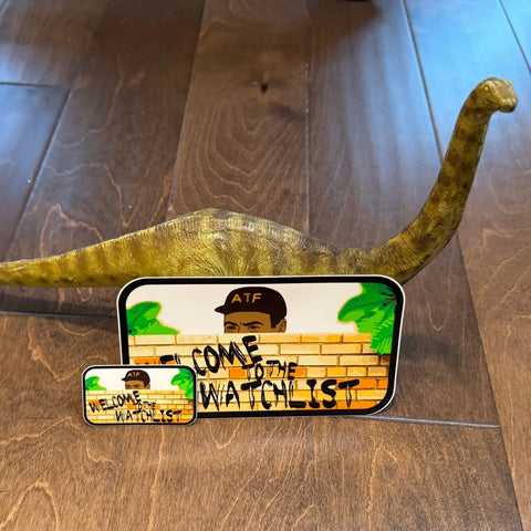 A toy dinosaur stands beside two American Lictor “Sticker: Welcome to the Watchlist” signs, each depicting a person in an ATF hat behind a graffiti wall, all set atop a wooden floor.
