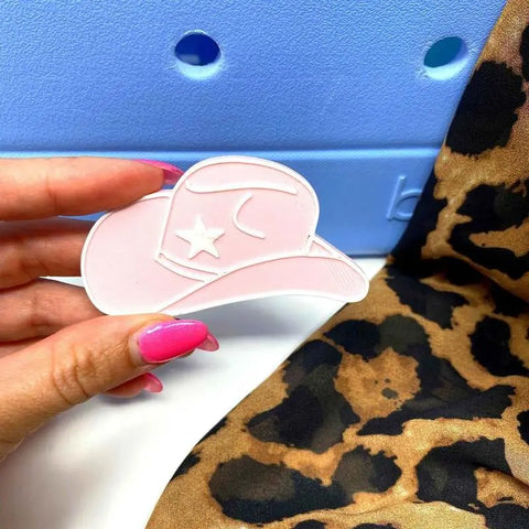 A hand with bright pink nails holds a pink cowboy hat-shaped BOGLETS - RODEO Charm by Chatelet Manufacturing—perfect as a decorative tote bag accessory. The background features light blue texture and brown and black leopard print fabric.