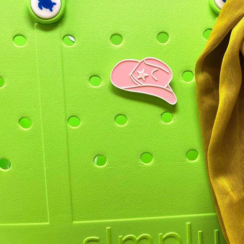 A lime green surface with circular holes displays a pink Chatelet Manufacturing BOGLETS - RODEO Charm with a star and yellow drape, plus two blue-and-white round Bogg Bag Accessories in the top left corner.