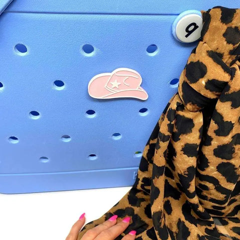 A hand with pink nails touches a blue perforated surface adorned with a pink BOGLETS - RODEO Charm by Chatelet Manufacturing, beside leopard print fabric—ideal for personalizing your tote with these unique, USA-made decorative bag charms.