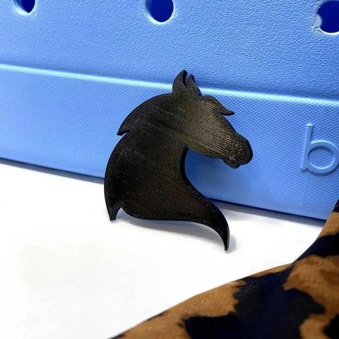 A BOGLETS - RODEO Charm by Chatelet Manufacturing, showing a black horse head silhouette, rests on a light blue tote bag with textured holes and embossed letters, while leopard print fabric appears in the corner. Made in USA.