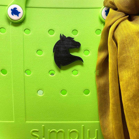 A BOGLETS - RODEO Charm by Chatelet Manufacturing, shaped like a black chess knight piece, decorates a bright green perforated tote with two white fasteners featuring blue designs and yellow fabric draped nearby. Made in USA.